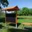 Outdoor Classroom - puinen ulkokatos, taulu ja puupenkit nurmikolla ympäröitynä puilla.